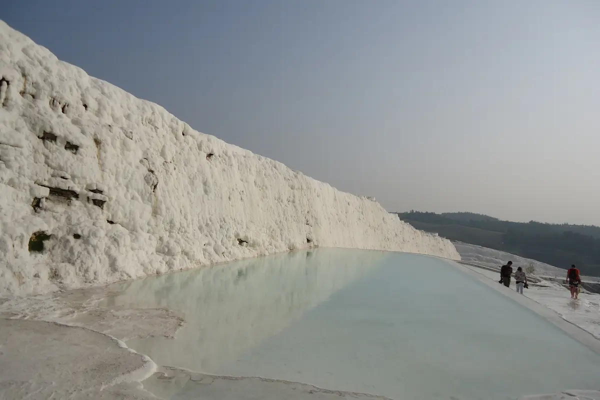 Pamukkale 一片白色方解石壁旁，一条淡蓝色钙华长池，三位游客赤脚沿着右侧白色小径行走